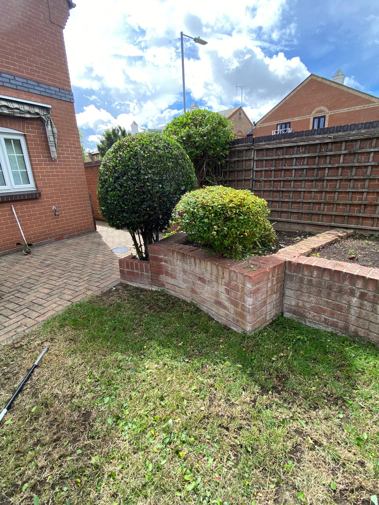 hedge trimming round shrubs beside low brick planter wall and paved path